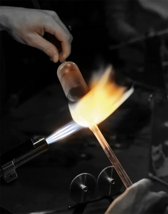 A close-up of a glassblowing process where a flame is being applied to a glass tube. The orange-yellow flame is heating the glass, causing it to glow as it is carefully shaped. A hand hovers nearby, controlling the process with precision. The tools and workspace are visible in the background, adding to the technical atmosphere of the craft.